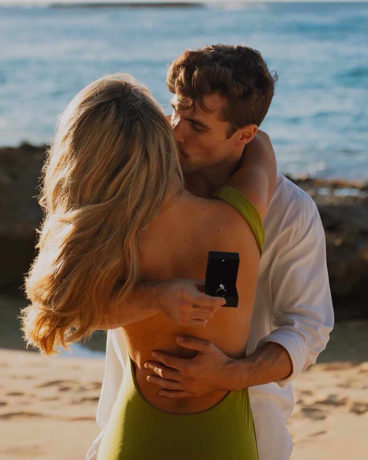A couple kisses on the beach after their Oahu proposal while he holds the open ring box