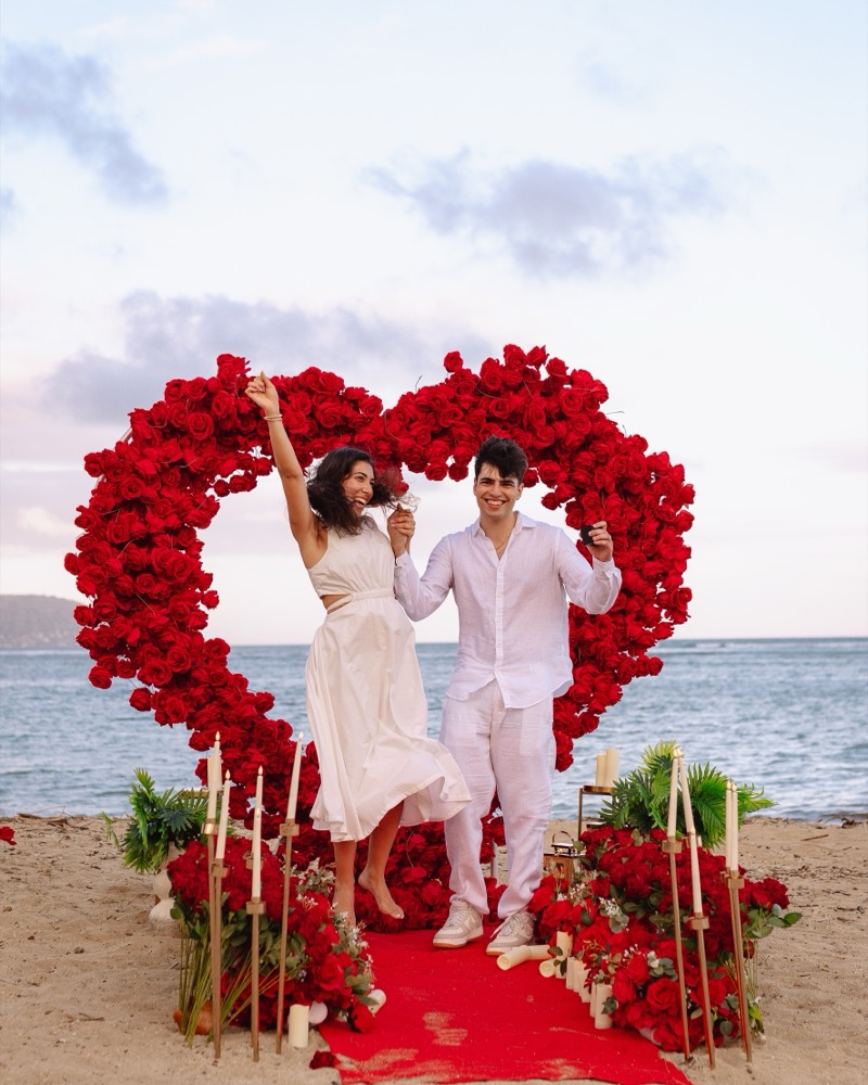 A couple celebrates their engagement in front of a red giant flower heart at Kahala Beach on Oahu