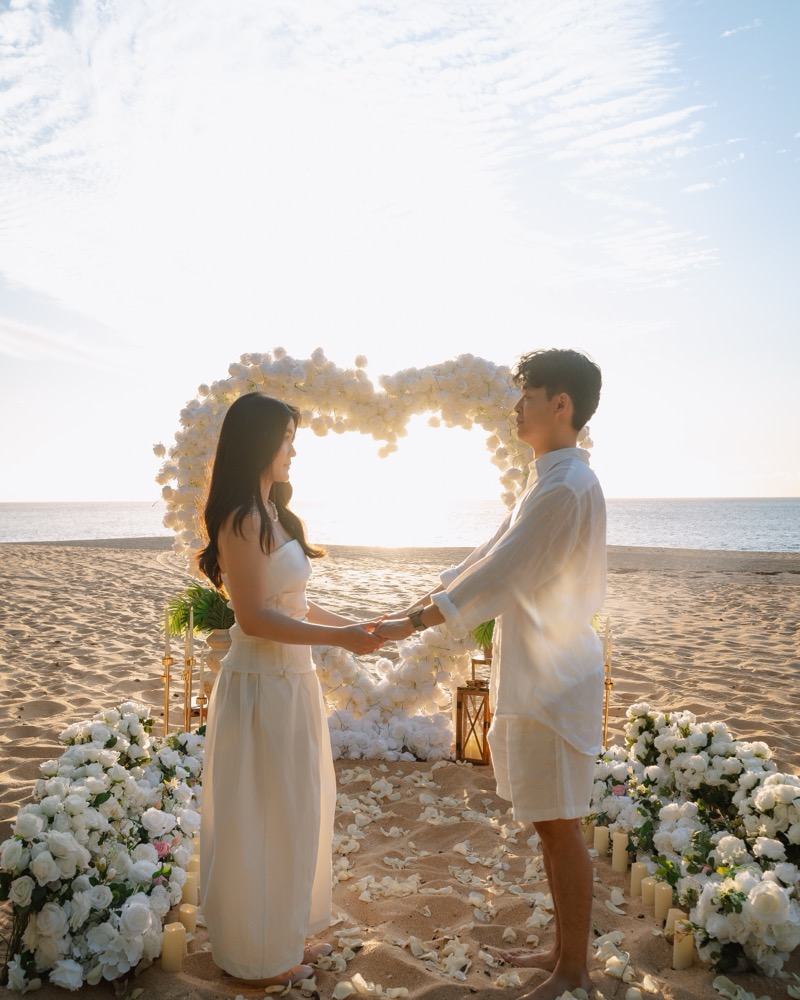 A couple holds hands in front of a white giant flower heart at sunset at Keiki Beach on Oahu