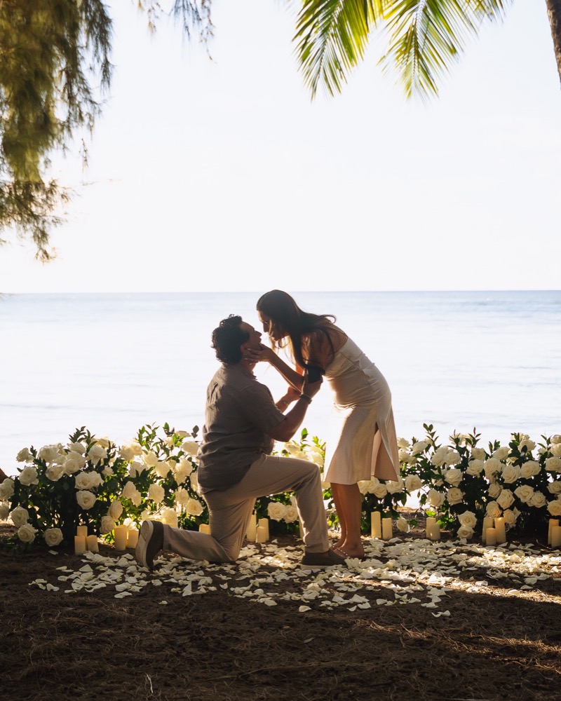 He proposes on one knee in front of white roses and candles under the palms at Kawela Bay on Oahu